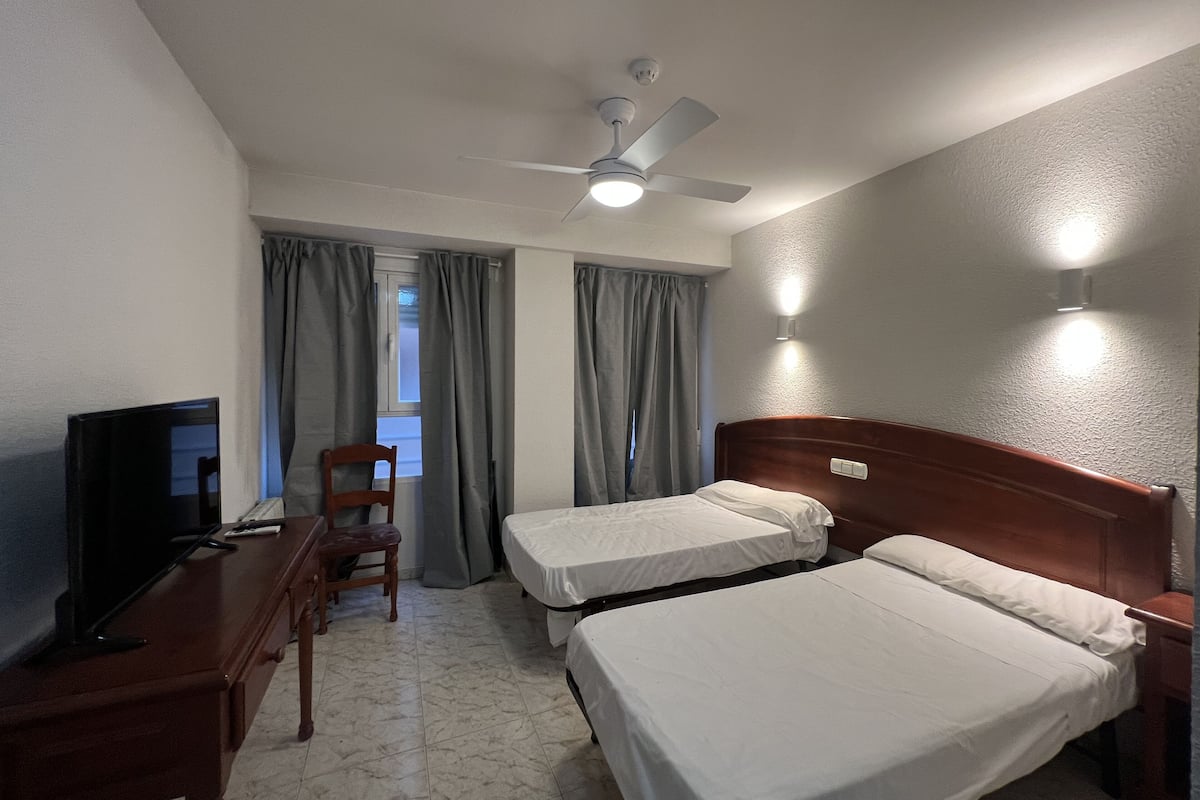 A hotel room with two beds, a desk with a television, a chair, a ceiling fan, and a window with curtains.