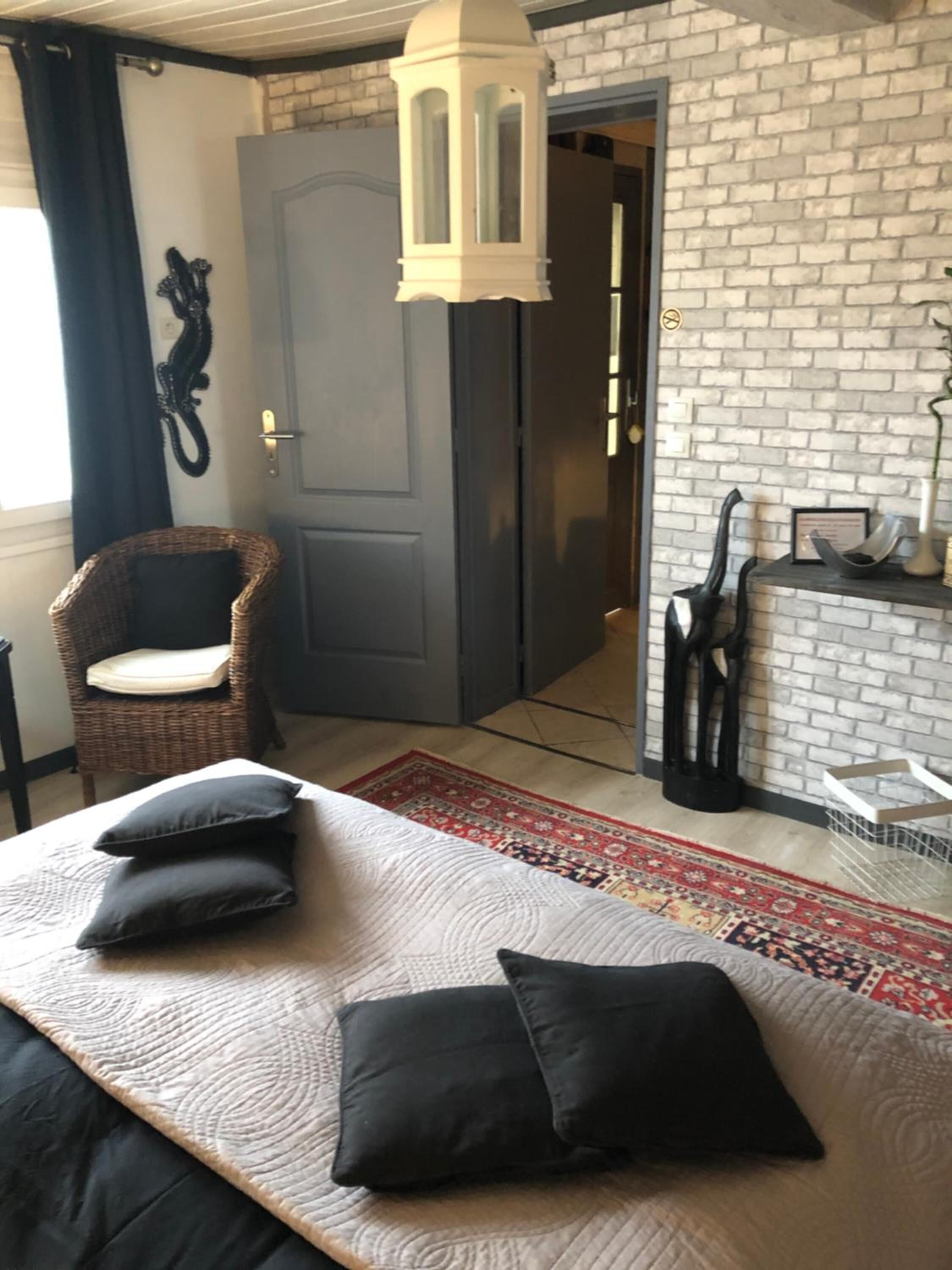 A room with a bed, a wicker chair, a door, a lamp, and a brick wall.
