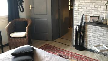 A room with a bed, a wicker chair, a door, a lamp, and a brick wall.