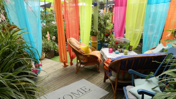 A cozy outdoor seating area with wicker chairs and a table, surrounded by colorful drapes and potted plants.