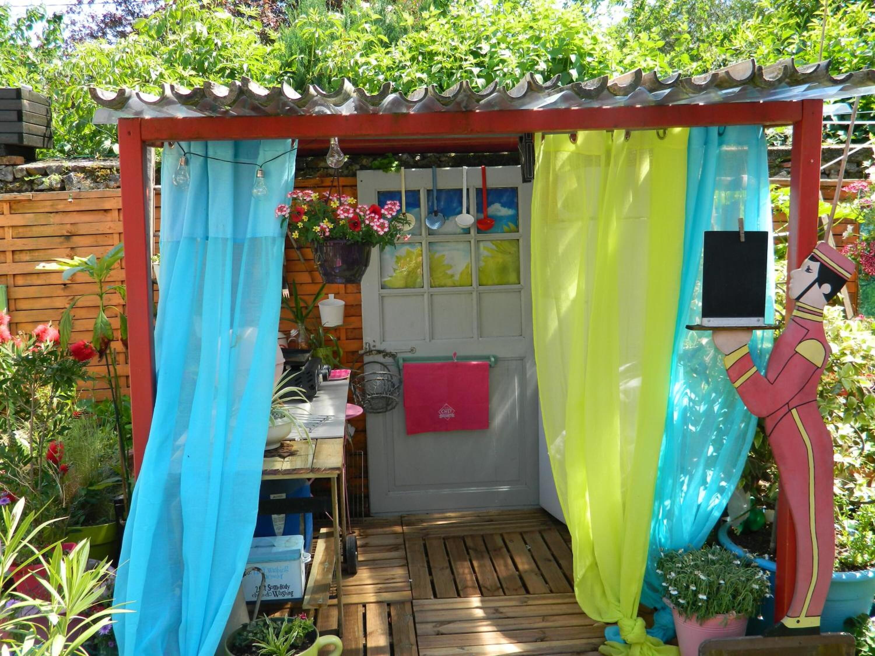 A small, enclosed garden area with a wooden structure, a window, and various plants and decorations.