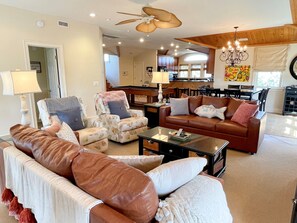 A spacious living room with a brown leather sofa, a patterned armchair, and a coffee table. A ceiling fan and a chandelier are visible. The room has a view of a kitchen and dining area in the background.