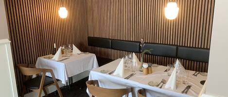 Un restaurante con dos mesas dispuestas para comer, cada una con mantel blanco, sillas negras y un muro de listones de madera al fondo.