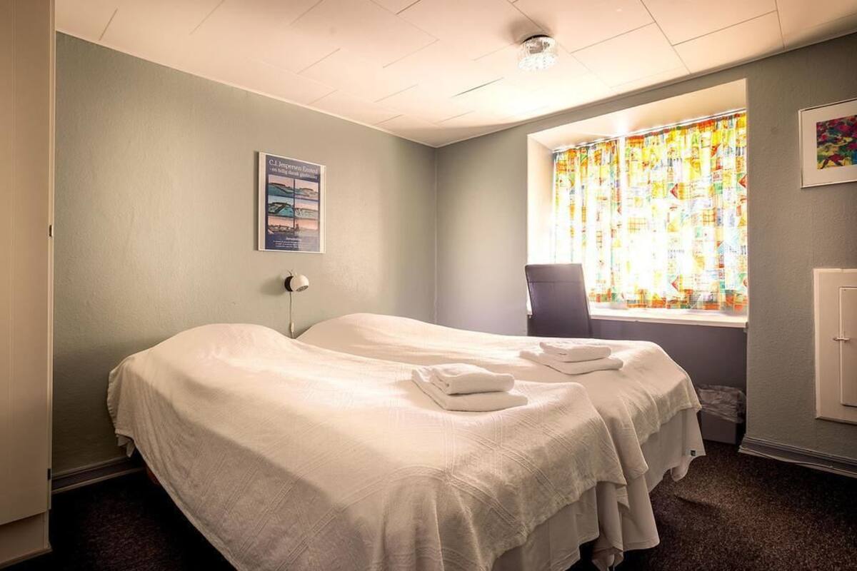 A hotel room with two beds, a window with colorful curtains, a poster on the wall, and a chair.