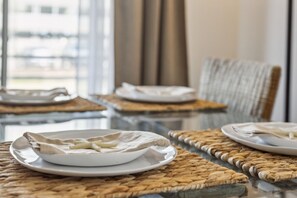 A dining table with plates and cutlery, set on woven placemats, in a room with a view through large windows. - Walk to beach, Patio, Pool, Central Downtown Condo (Virginia Beach)