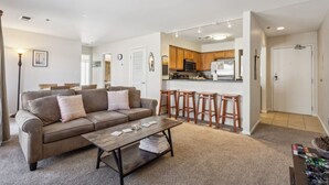 Sala de estar con sofá, mesa de centro y zona de cocina con taburetes de madera y refrigerador.