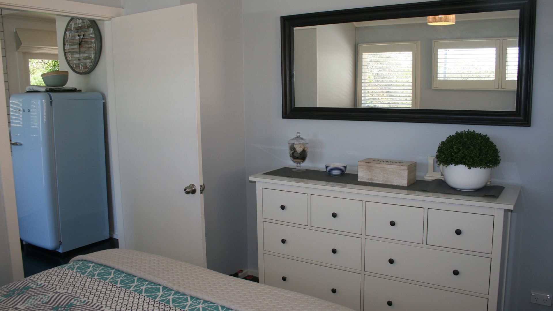 A bedroom with a bed, a dresser with drawers, a mirror, a clock, and a window with blinds.