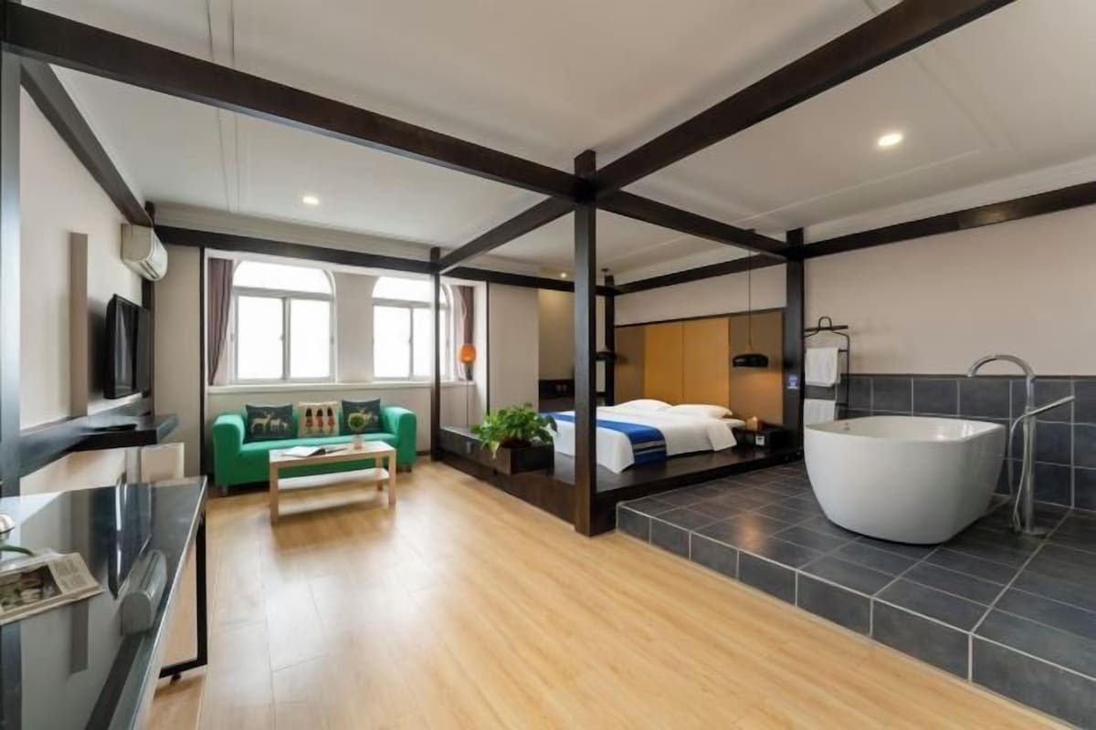 A modern hotel room with a large bed, a green sofa, a small table, a TV, and a large bathtub.