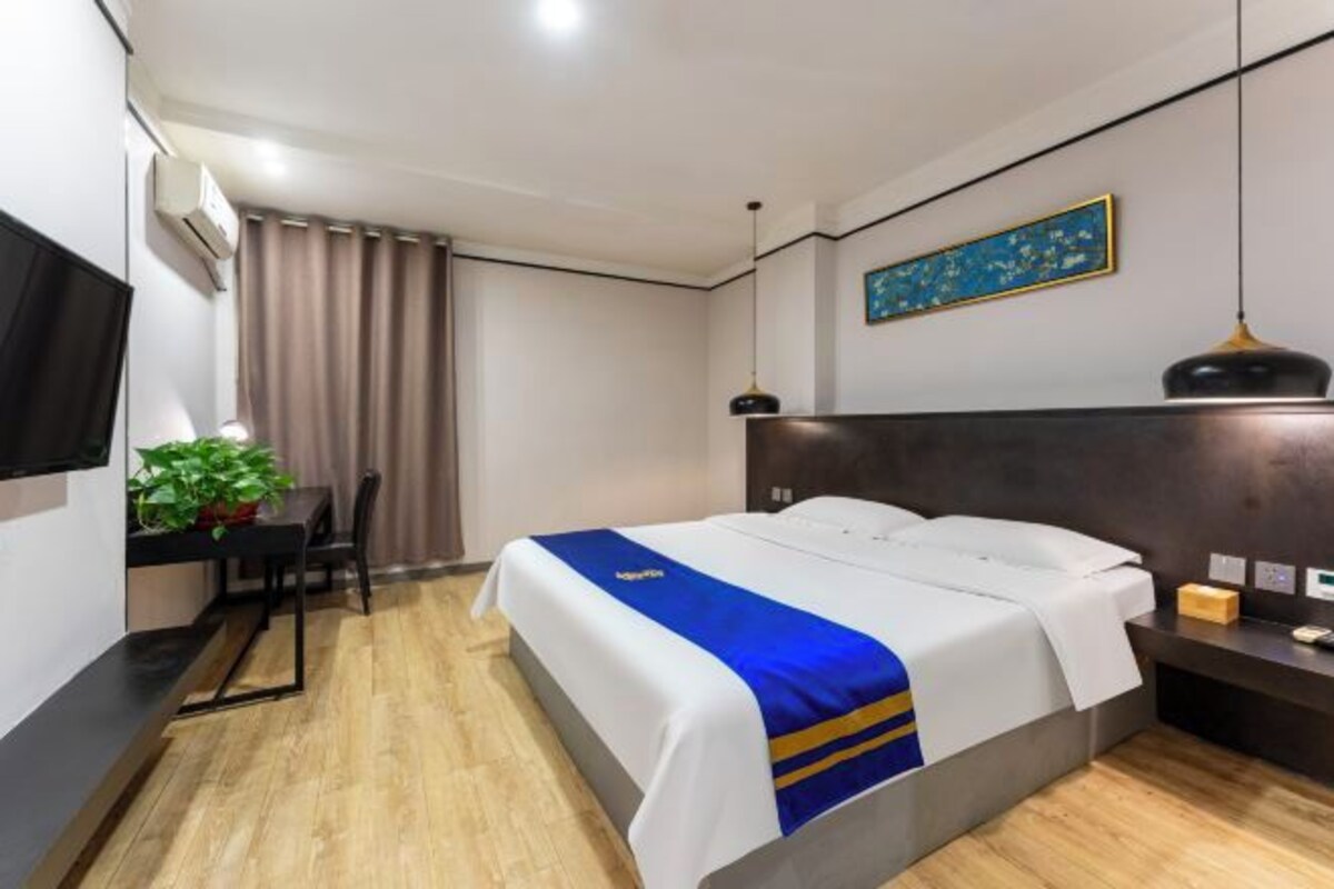 A hotel room with a bed, a desk, a chair, a potted plant, and a painting on the wall.