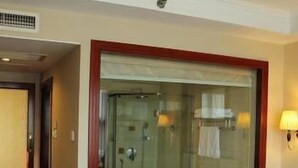Ein Hotelzimmer mit einer großen Duschkabine aus Glas, einem Bett mit weißer Bettdecke sowie einem Nachttisch mit Lampe und Telefon.