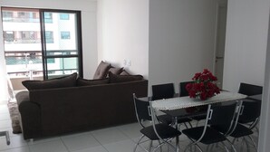 A living room with a brown sofa, a dining table with chairs, and a vase of red flowers. - Para 9 Pessoas ao Lado Shopping Rio Mar (Fortaleza)