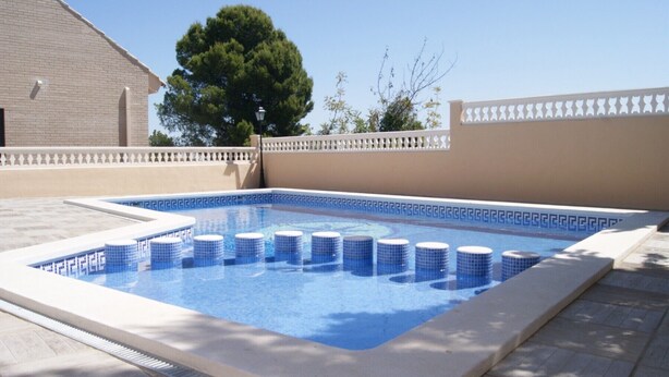 Una piscina rectangular con azulejos azules y escalones blancos, rodeada por un muro beige con detalles blancos decorativos.