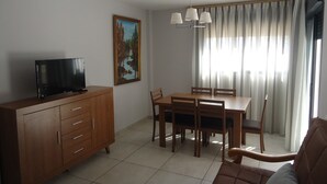 Una sala de estar con un televisor sobre un mueble de madera, una mesa de comedor con sillas, un cuadro en la pared y una ventana con cortinas.