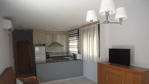 Cocina compacta con refrigerador, estufa y cafetera. Hay un mueble de madera con un televisor encima. Un sofá con cojines está en primer plano.
