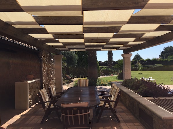 Ein überdachter Patio-Bereich mit Holztisch und -stühlen, Steinmauern und Blick auf einen angelegten Garten.