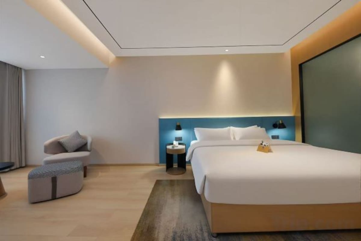 A modern hotel room with a large bed, a seating area with a chair and ottoman, a bedside table with a lamp, and a large window.