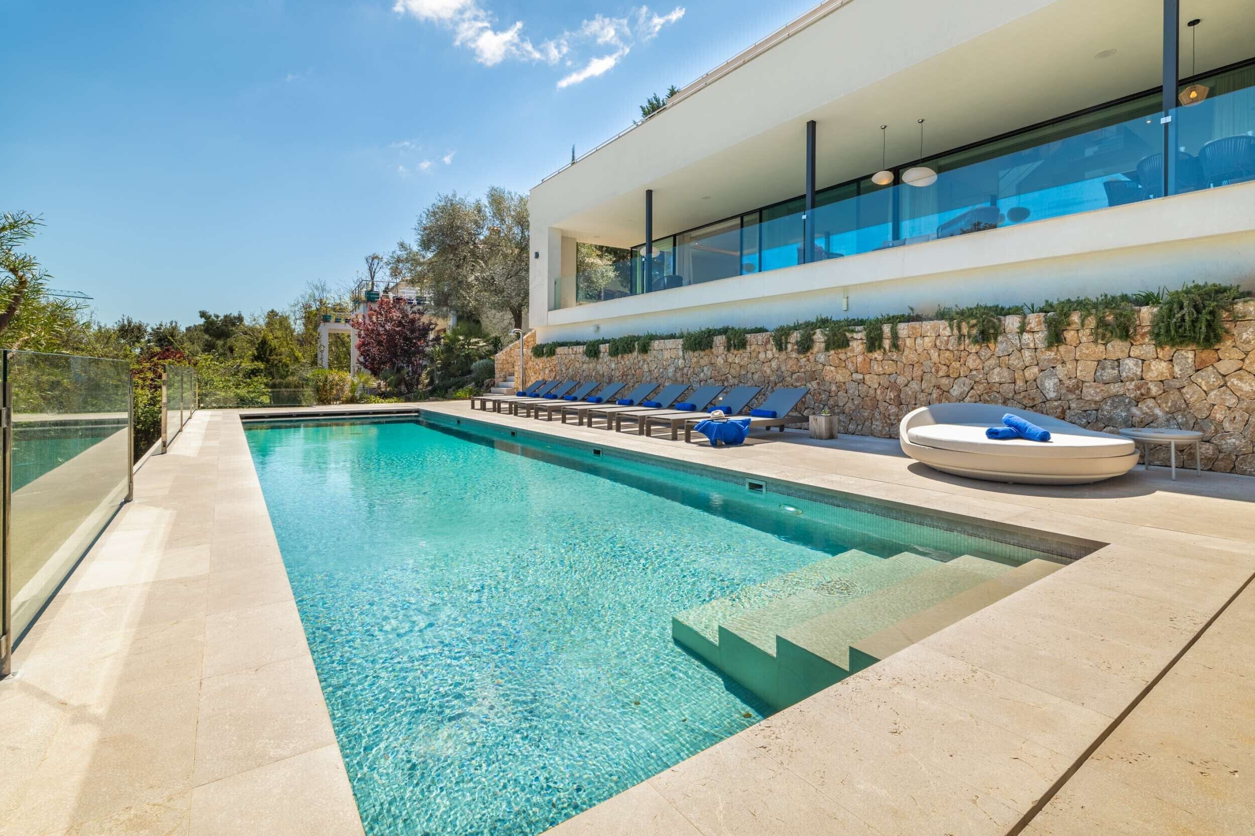 A modern pool area with clear blue water, sun loungers, and a stone wall.