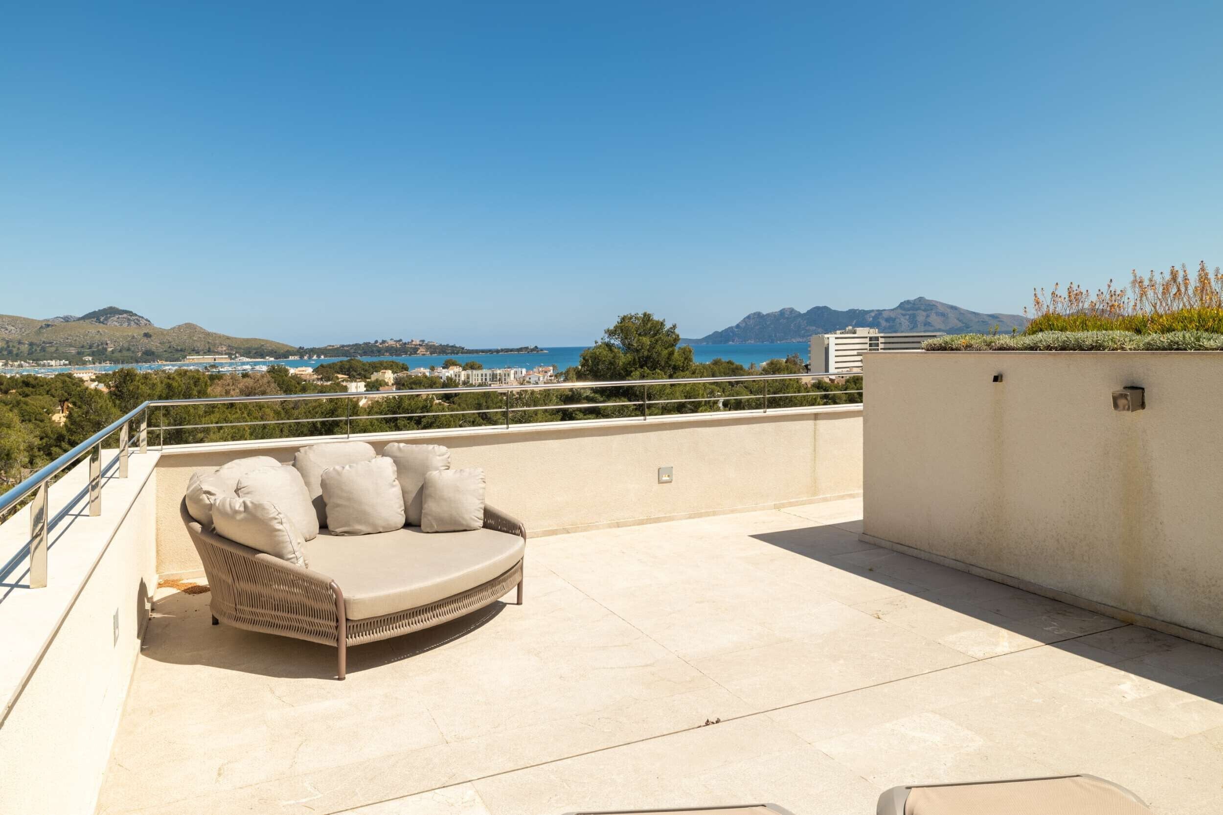 A rooftop terrace with a beige sofa and cushions, a railing, and a view of the sea and mountains.