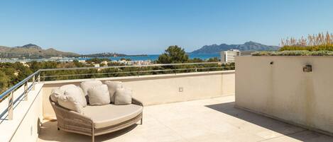A rooftop terrace with a beige sofa and cushions, a railing, and a view of the sea and mountains.