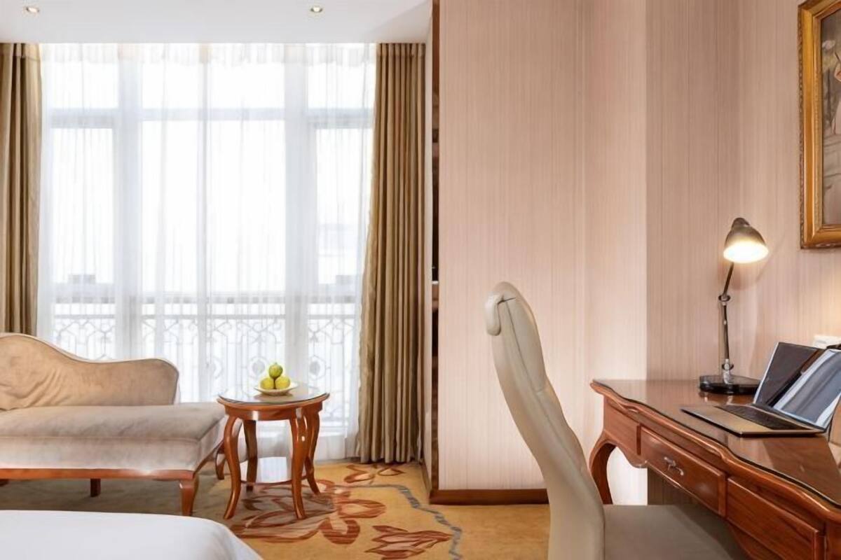 Habitación de hotel con cama, escritorio, silla, una mesita con fruta y ventana con cortinas.