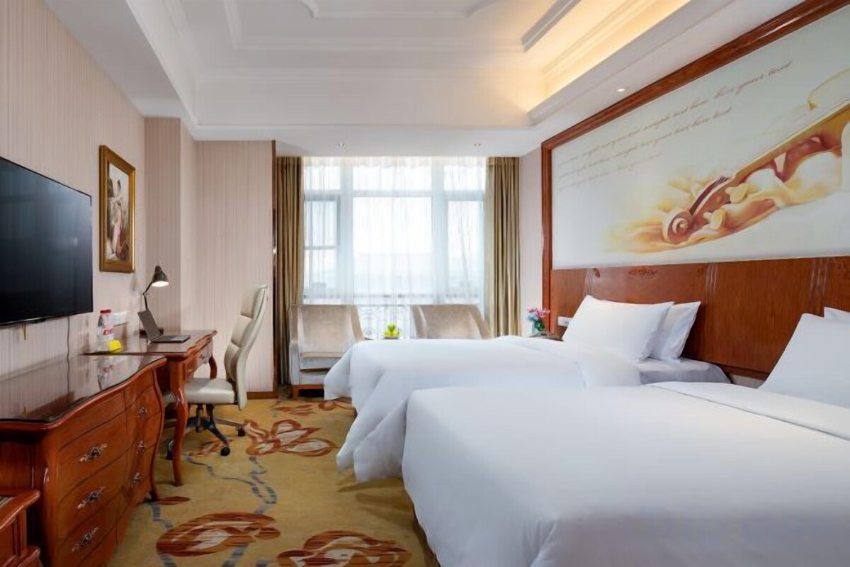 A hotel room with two beds, a desk with a chair, a television, and a large window with curtains.