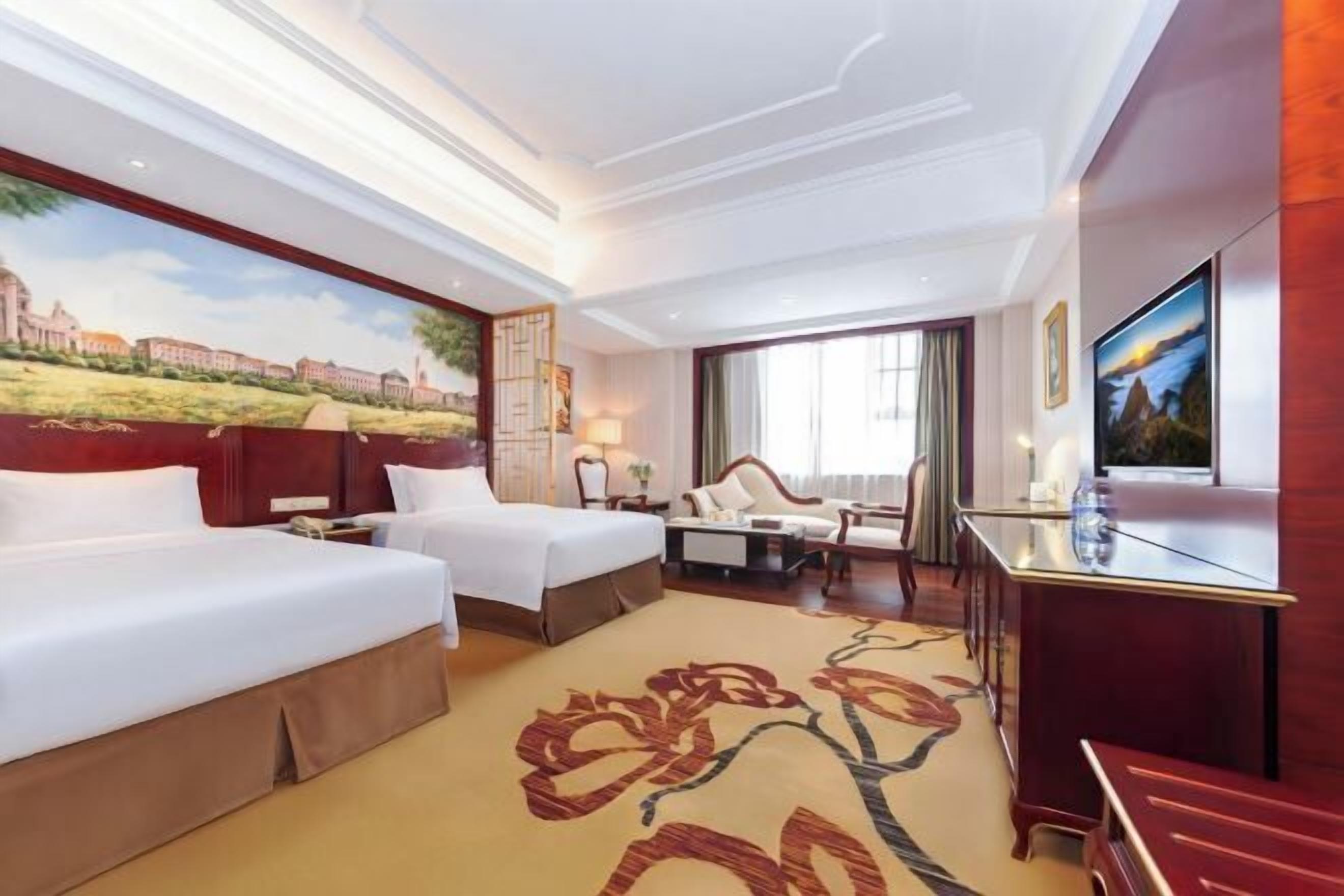 A hotel room with two beds, a desk, a TV, and a large window.