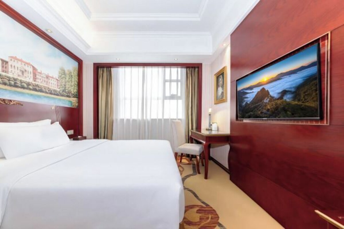 A hotel room with a bed, a desk, a chair, a painting on the wall, and a window with curtains.
