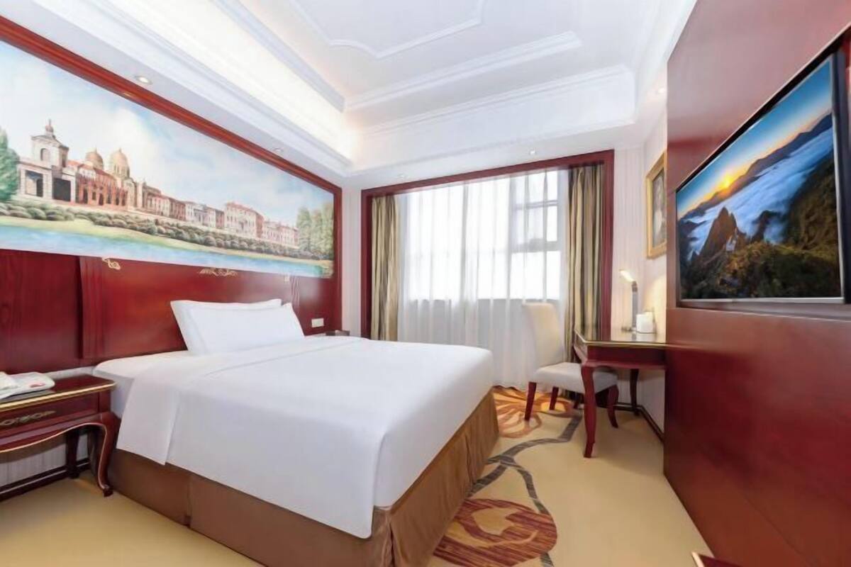 Quarto de hotel com uma cama grande, uma escrivaninha, uma cadeira, um quadro na parede e uma janela com cortinas.