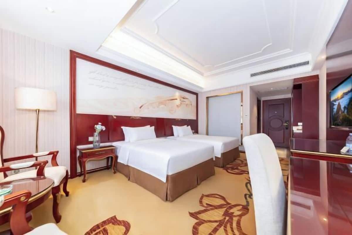 Quarto de hotel com duas camas, uma escrivaninha, uma cadeira, um abajur e um quadro na parede.