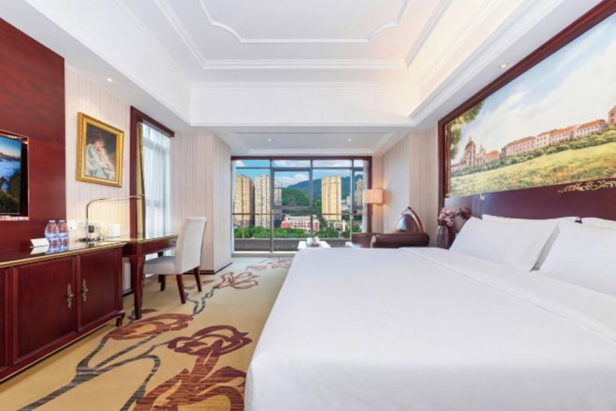 A hotel room with a large bed, a desk, a chair, a TV, and a window with a cityscape view.