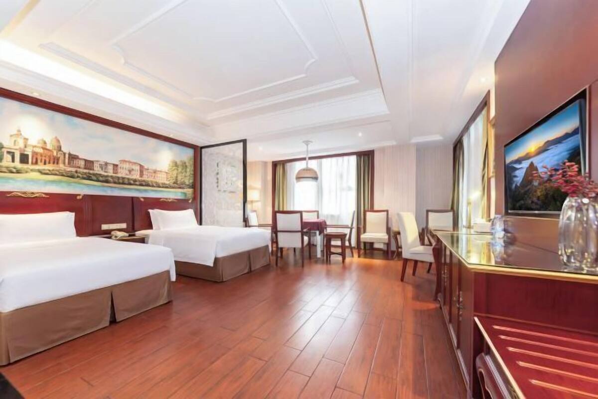 Quarto de hotel com duas camas, uma escrivaninha, uma cadeira, uma televisão e um quadro na parede.