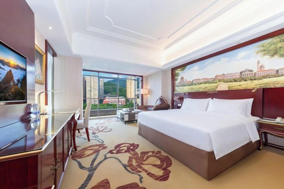A hotel room with a large bed, a desk with a chair, a television, and a large window offering a city view.