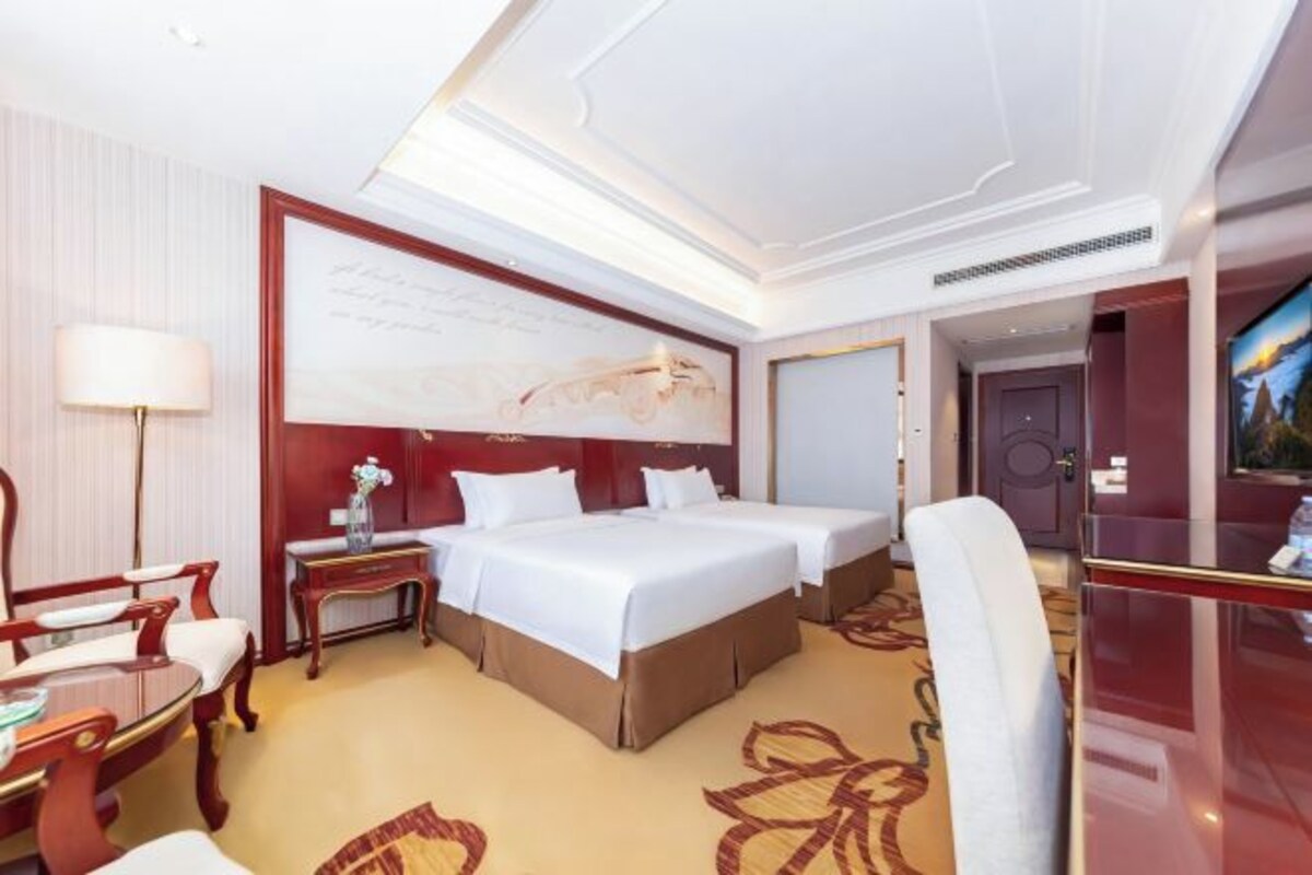 A hotel room with two beds, a desk, a chair, a lamp, and a painting on the wall.