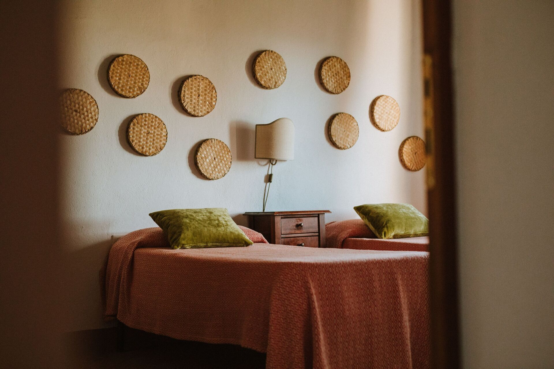 A room with two beds, each with a green pillow, a small wooden nightstand, and a wall-mounted lamp.