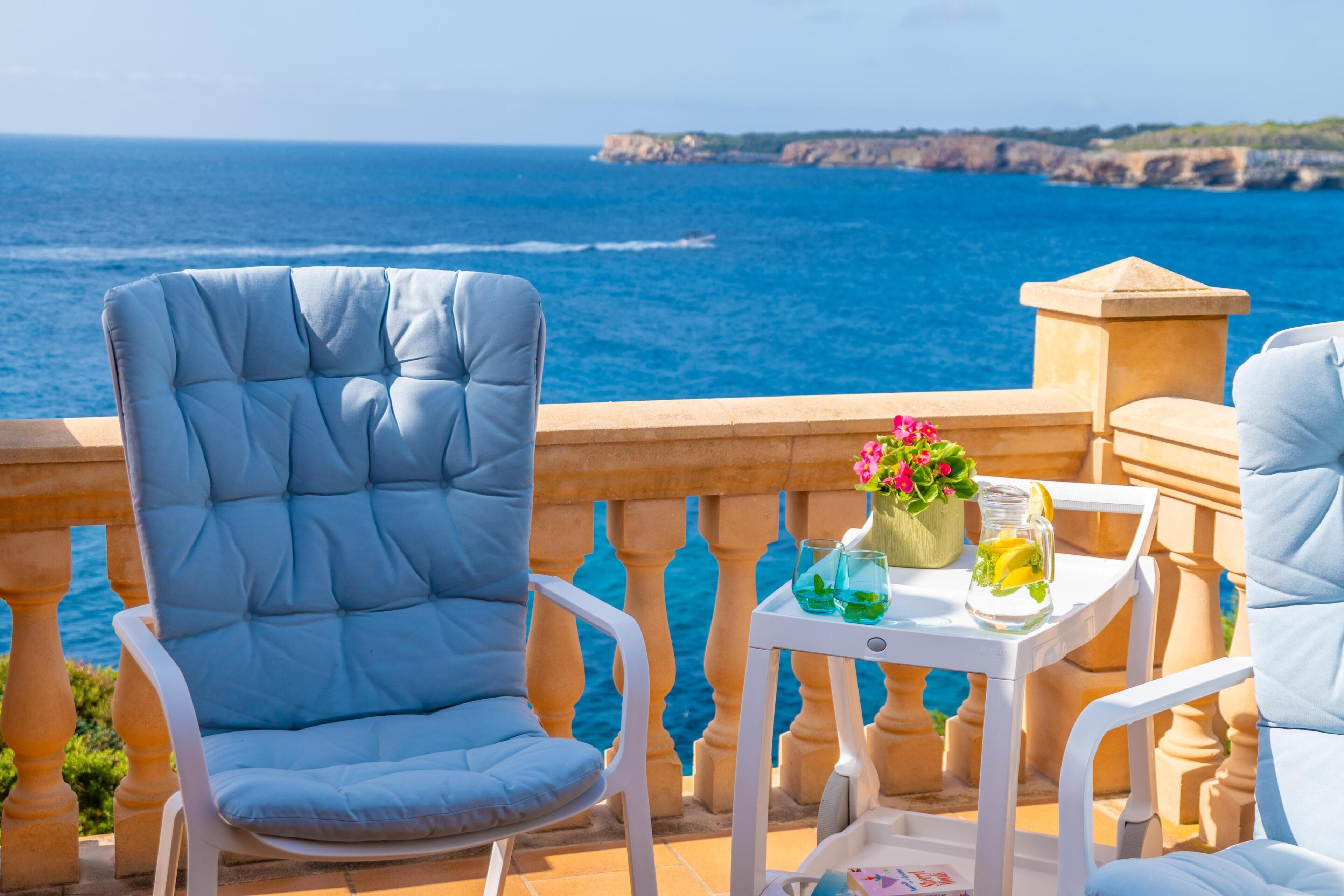 Een balkon met uitzicht op de zee, met twee blauwe stoelen met kussens en een kleine tafel met een bloemenvaas en een glas limonade.