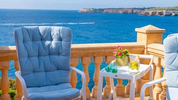 Een balkon met uitzicht op de zee, met twee blauwe stoelen met kussens en een kleine tafel met een bloemenvaas en een glas limonade.