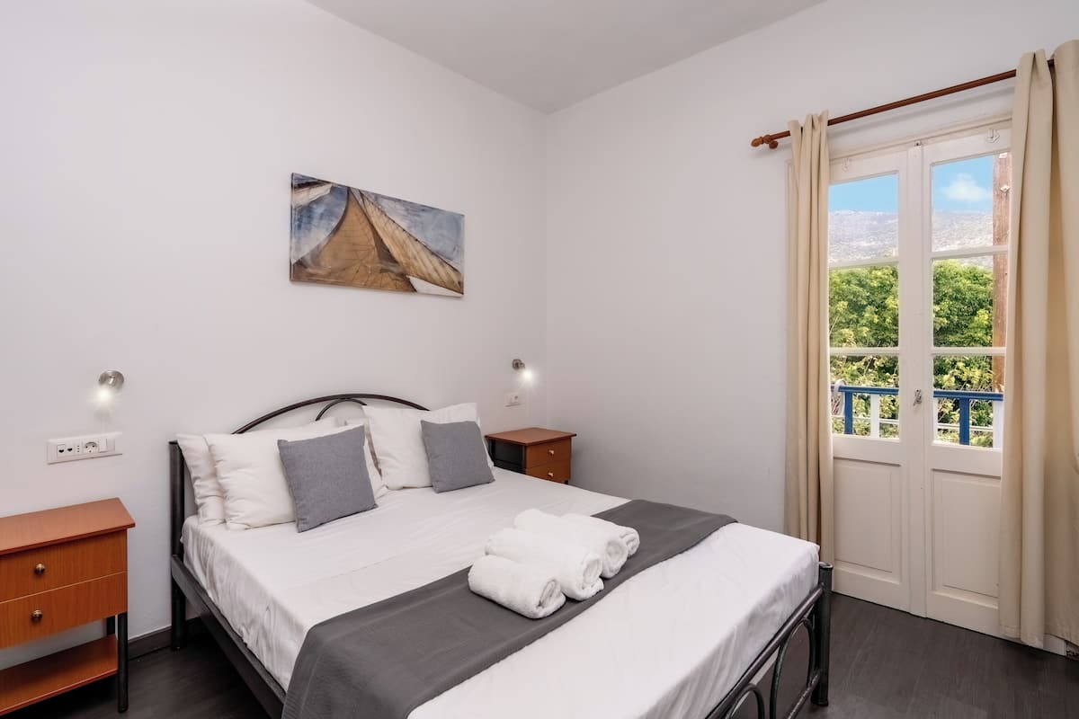 A neatly made bed with white linens and a gray throw, a painting on the wall, and a window with curtains.