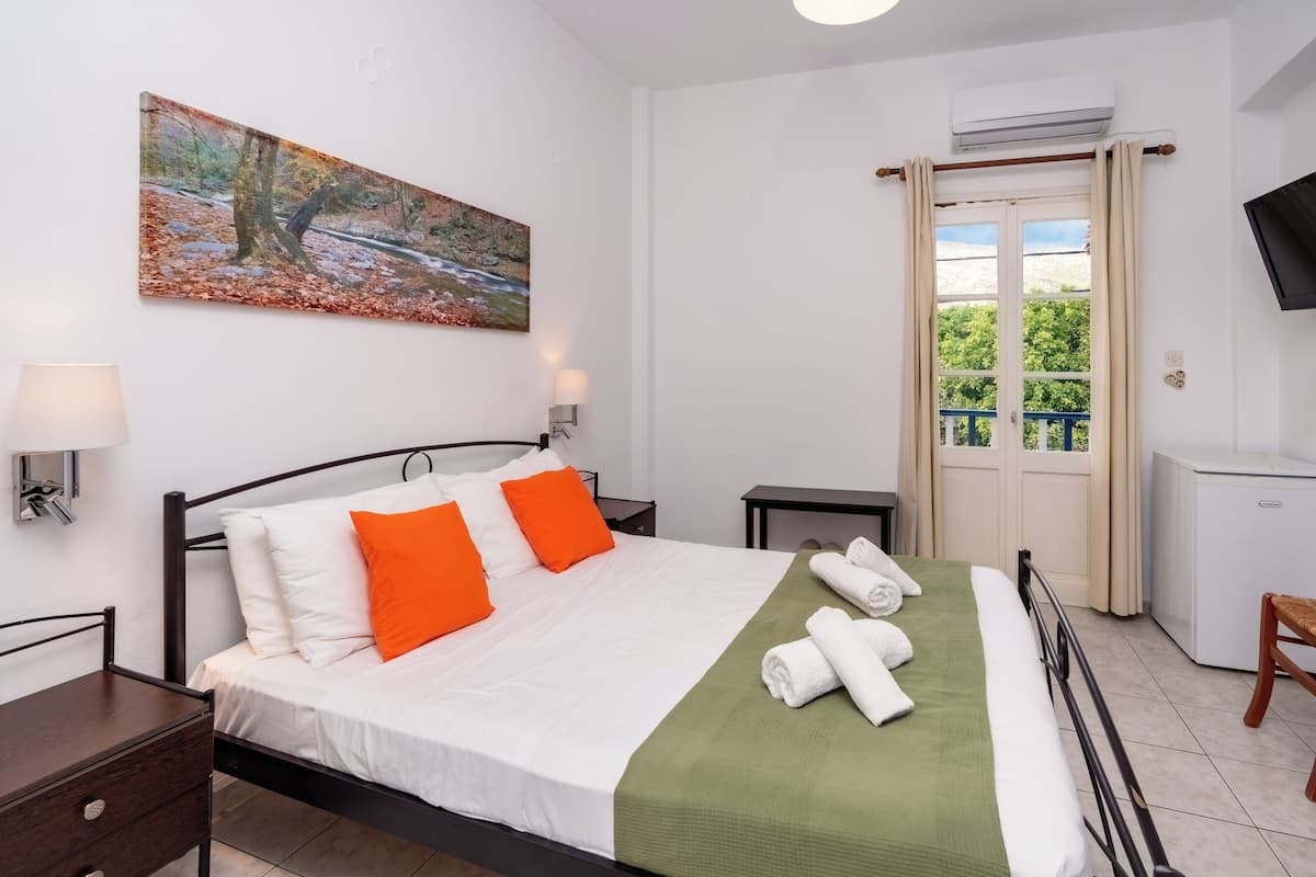 A neatly arranged bedroom with a bed, a bedside table, a lamp, a painting, an air conditioning unit, a window with curtains, and a television.