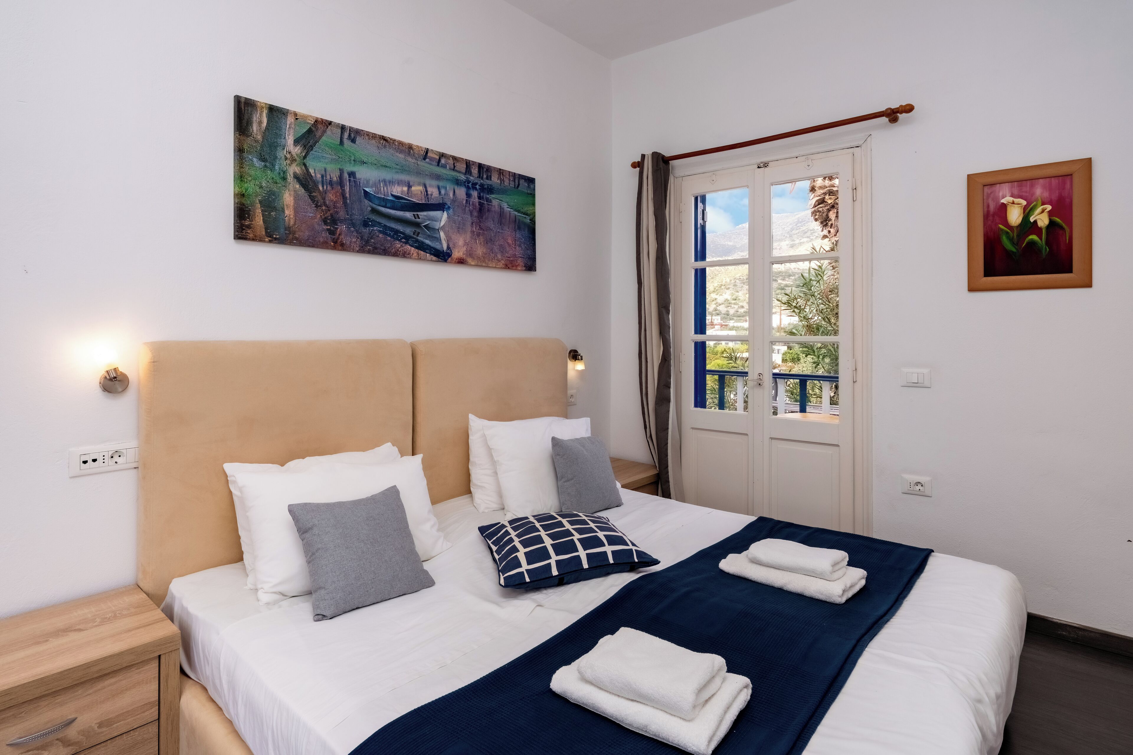 A neatly made bed with white and gray pillows, a blue blanket, and folded towels. There is a painting of a landscape on the wall and a framed picture of flowers.