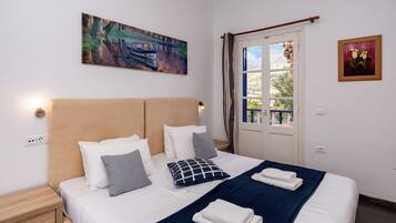 A neatly made bed with white and gray pillows, a blue blanket, and folded towels. There is a painting of a landscape on the wall and a framed picture of flowers.