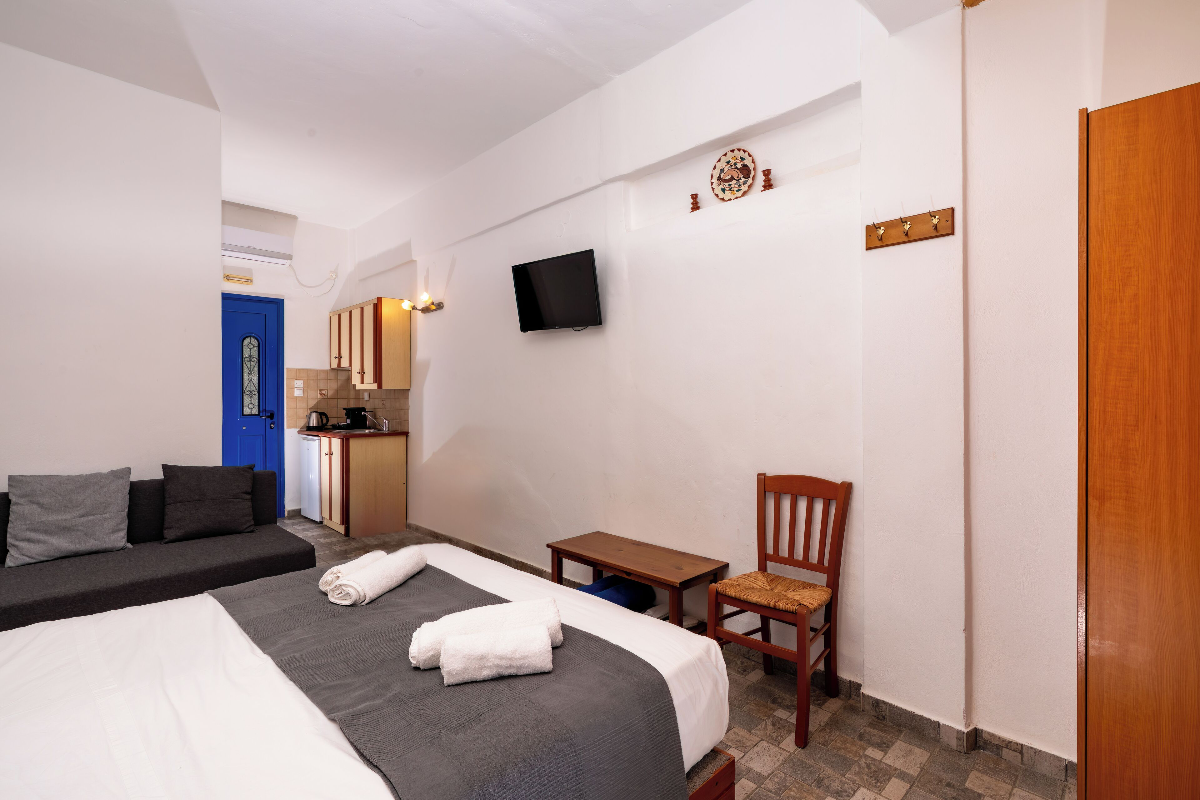 A hotel room with a bed, a chair, a small table, a TV, and a kitchenette in the background.