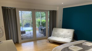 Une chambre d’hôtel moderne dotée d’une grande porte-fenêtre coulissante donnant sur un patio extérieur, d’un canapé blanc, d’un lit avec des serviettes soigneusement pliées et d’un plancher en bois.