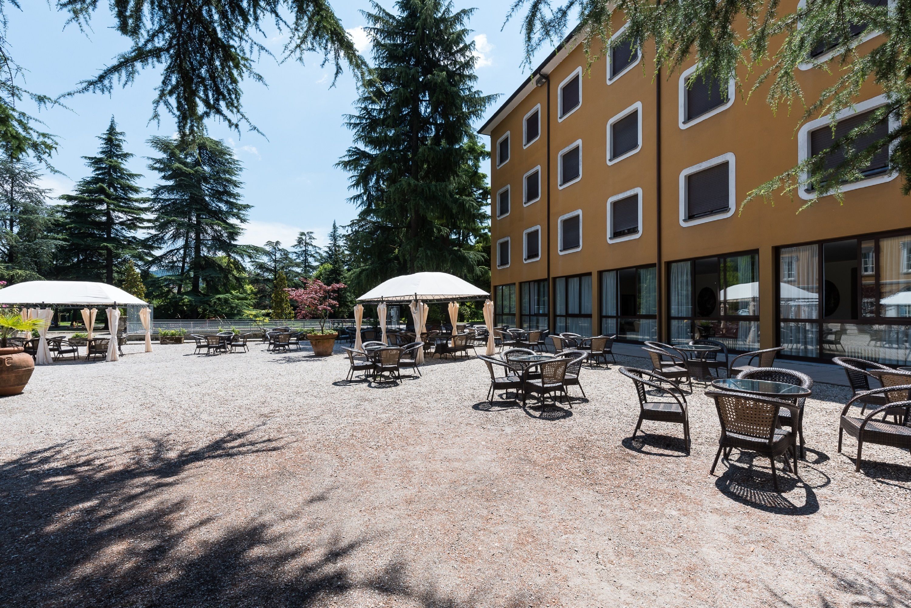 An outdoor seating area with tables and chairs, surrounded by trees and a building with large windows.