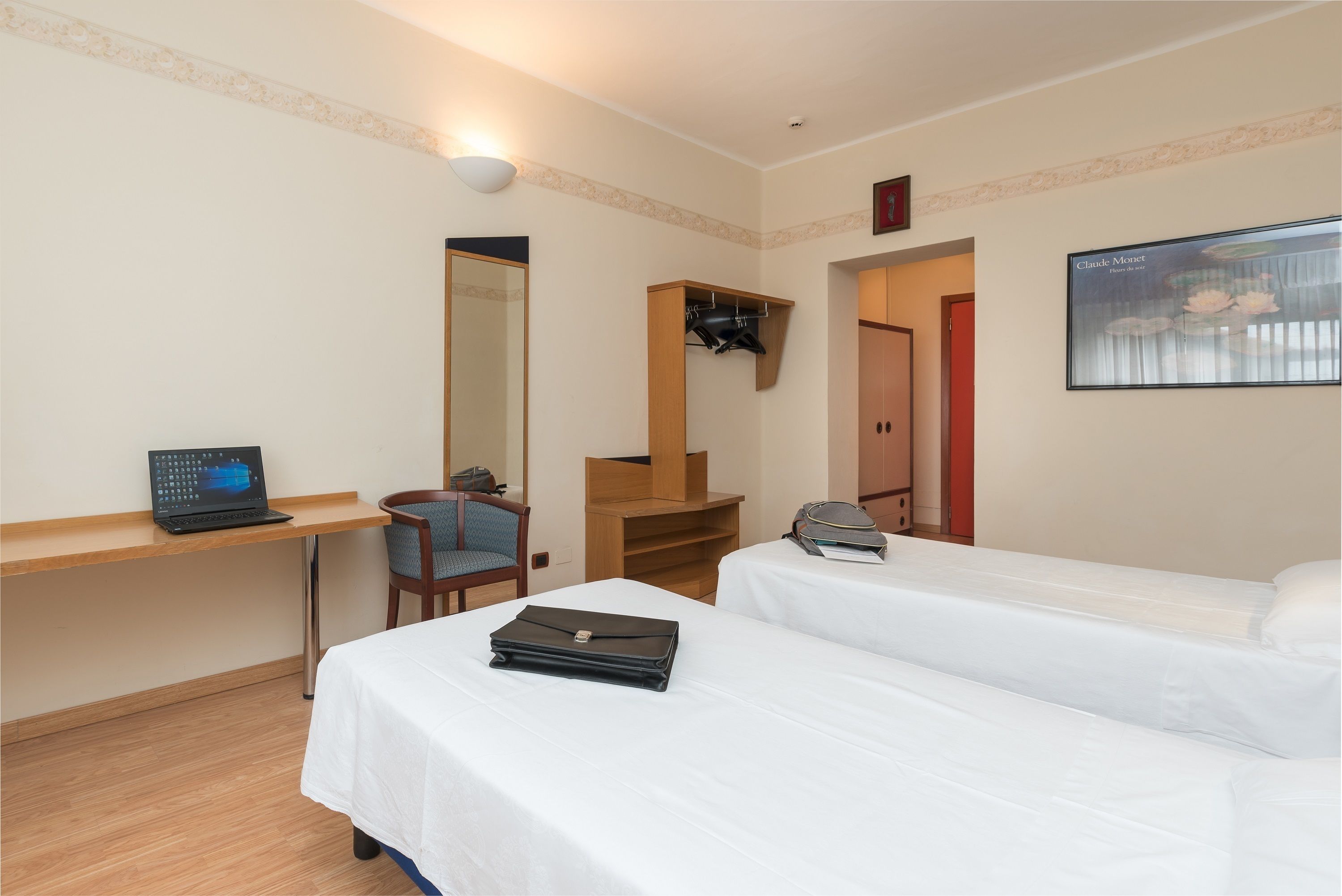 A hotel room with two beds, a desk with a laptop, a chair, a mirror, a coat rack, and a framed picture on the wall.