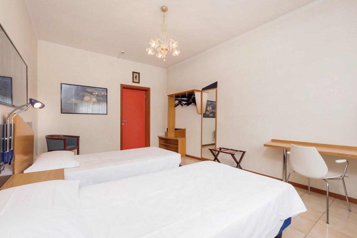 Une chambre d’hôtel avec deux lits, un bureau, une chaise, un miroir, une porte rouge et un lustre.