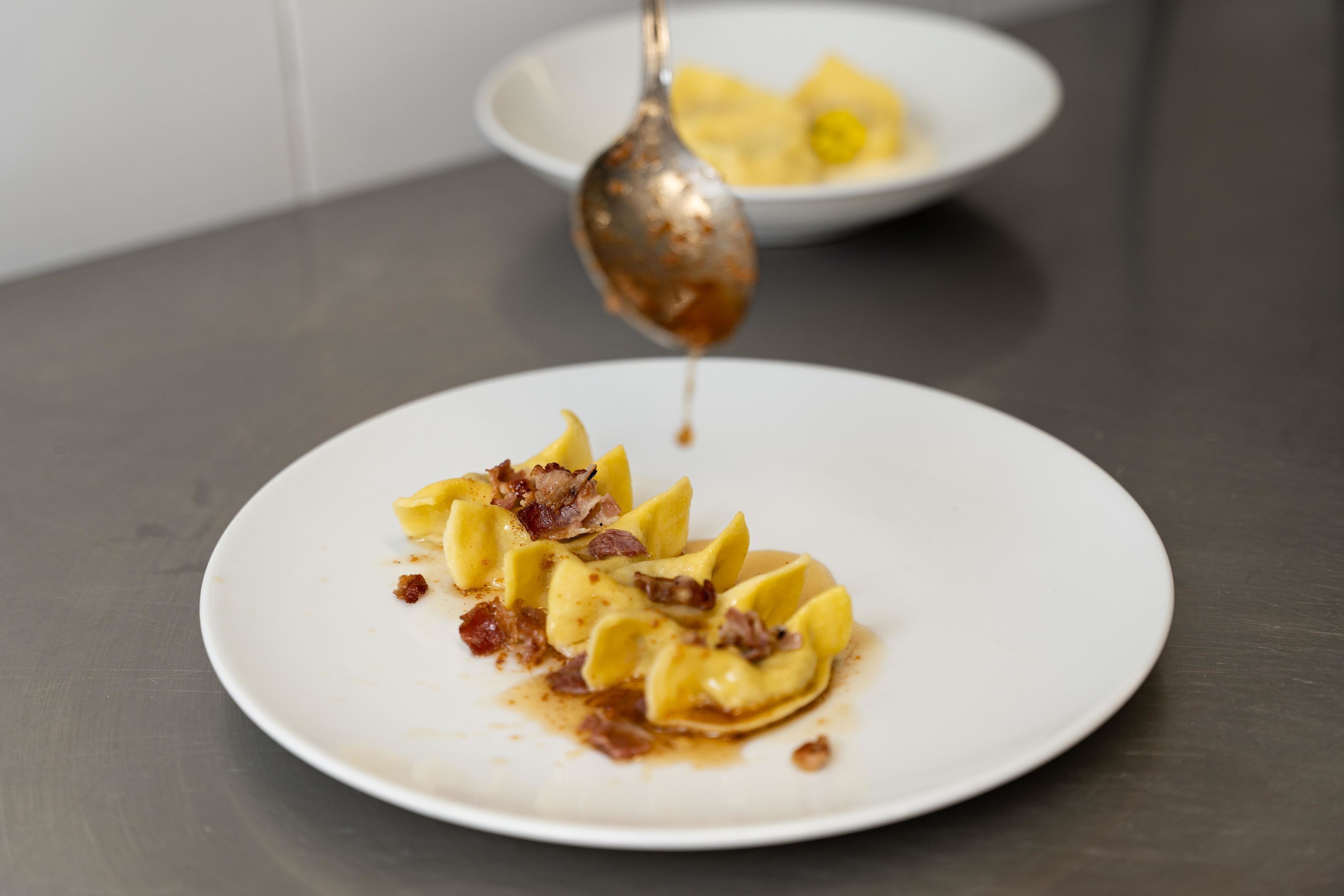 Een wit bord met ravioli, overgoten met saus en spek, en een lepel die saus opschept.