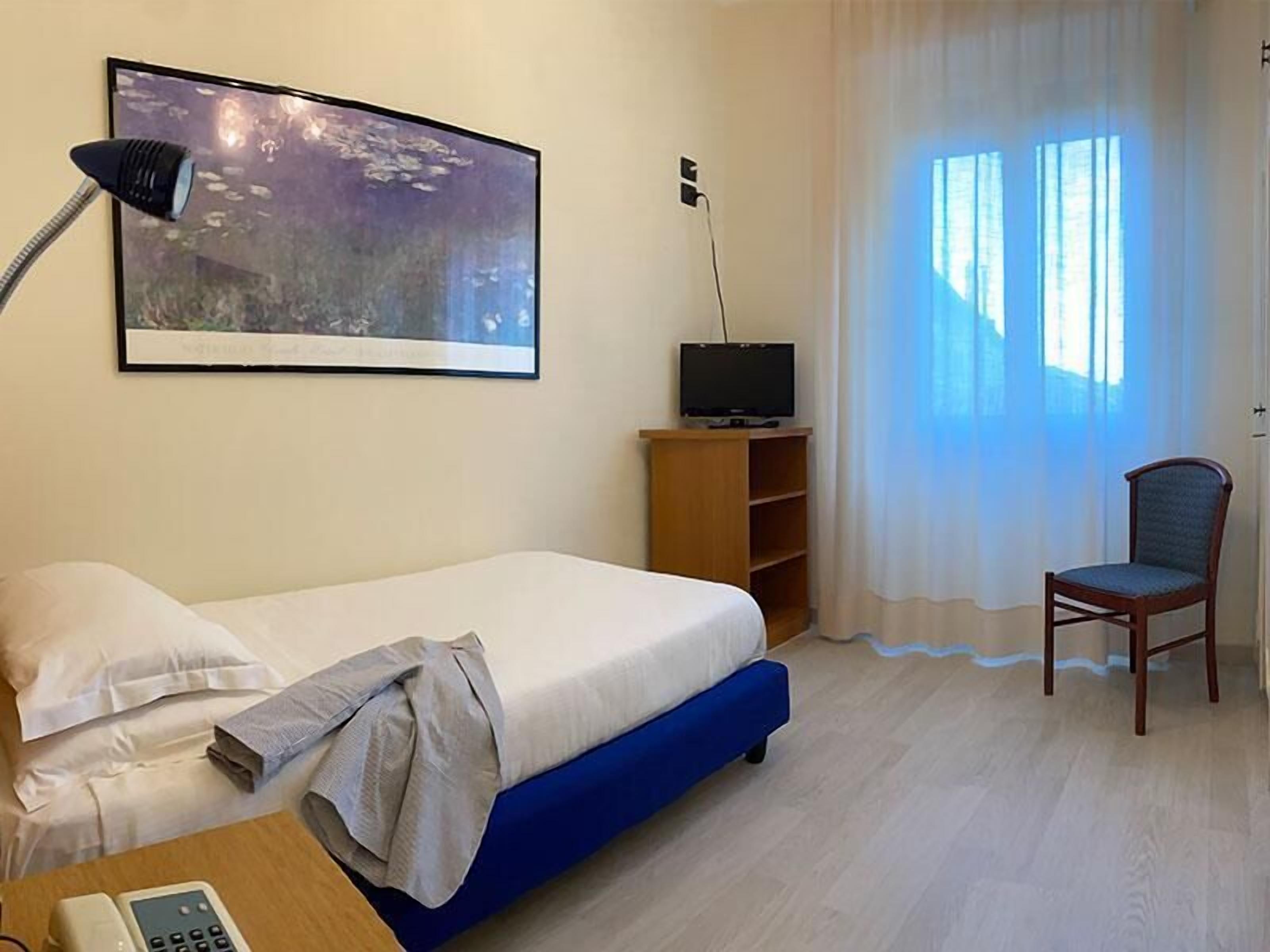 A hotel room with a bed, a desk with a telephone, a television on a wooden stand, a chair, and a large window with curtains.