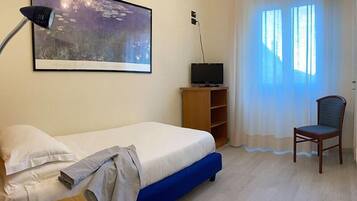 A hotel room with a bed, a desk with a telephone, a television on a wooden stand, a chair, and a large window with curtains.
