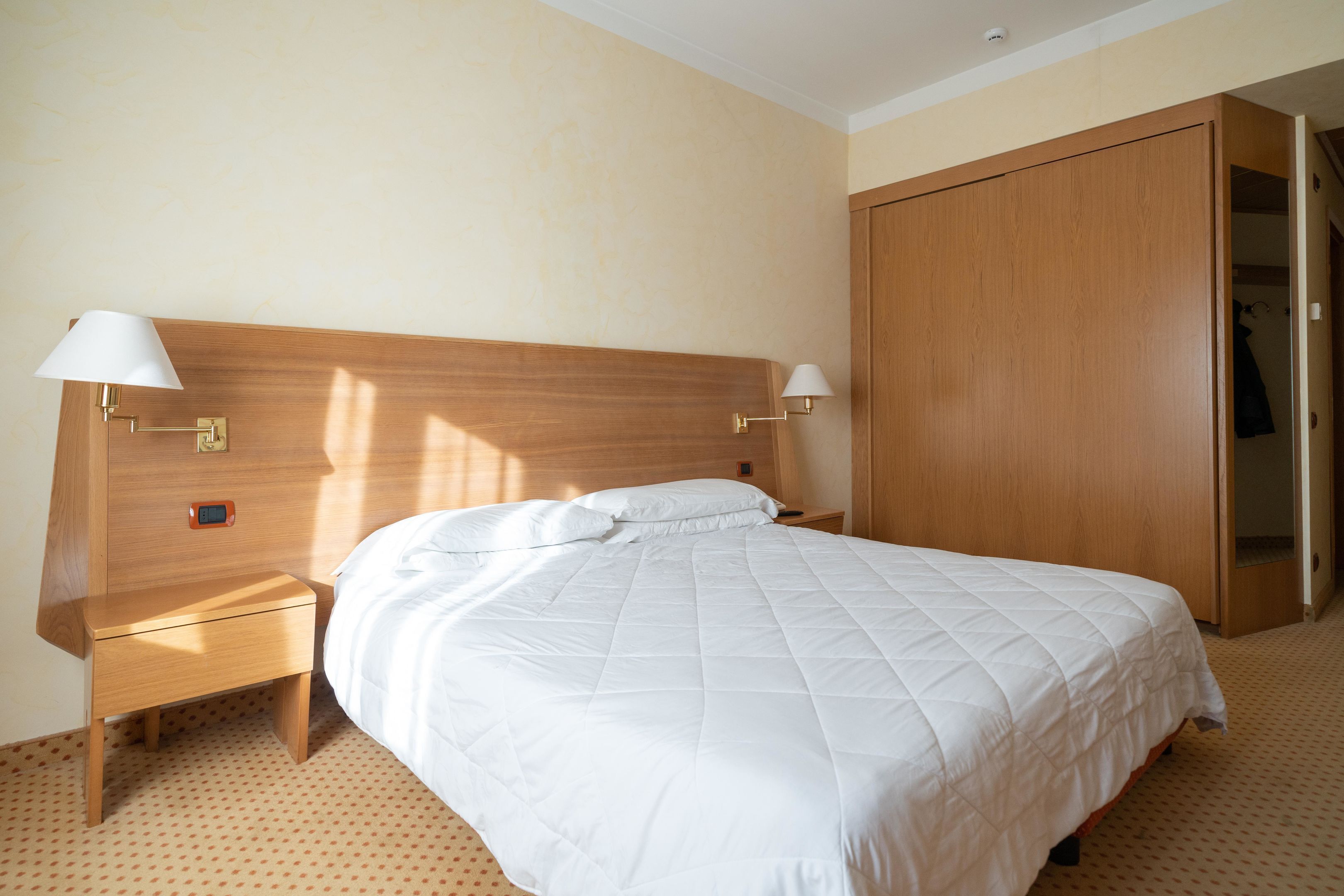 A hotel room with a wooden headboard, a white bedspread, two bedside lamps, a nightstand, and a wardrobe.