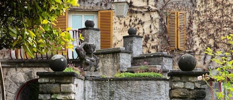 A stone staircase leading to a building with wooden shutters, surrounded by greenery and decorative stone elements.
